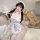 A woman poses with a teddy bear, wearing a white cherry-themed maid outfit featuring lace details and thigh-high stockings. t...