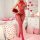 A woman stands in a heart-themed bedroom holding a bouquet of red roses, wearing a red valentines day bodysuit with sheer mes...