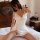 A woman poses elegantly on a bed wearing a white lace-backless sleep dress featuring a deep v-neckline and high slit, exuding...
