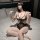 Woman in black sheer mesh lingerie set with lace trim and chain detail, sitting on bed.