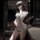 Woman wearing a sleek, form-fitting gray bodysuit with lace detailing, posing by a window at night — perfect for yoga or date...