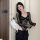 Woman modeling a black lace bodysuit with sheer sleeves and floral patterns, paired with black pants and a white handbag.