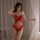 Woman wearing red lace deep v bodysuit with sheer mesh and open-crotch design, posing by white curtains.