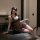 Woman wearing black lace transparent babydoll set with sheer details, posing seductively on table in dimly lit room.