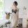 A woman stands elegantly holding a basket of flowers, wearing a beige silk dress with black lace detailing. the setting featu...